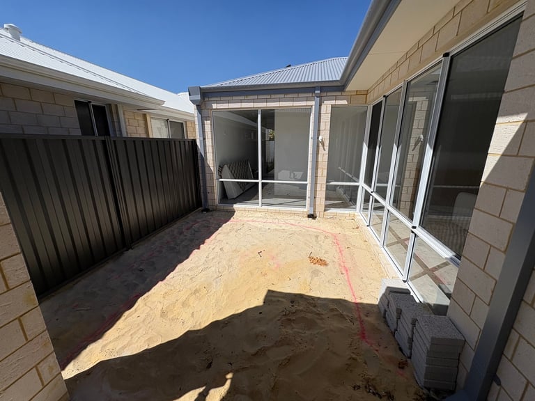 Backyard patio area under construction with sand, concrete blocks, and a modern house with glass doors and metal fencing